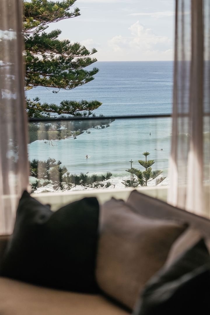 Wellness Suite King at Elysium Manly with pillows framing views of pine trees and surfers floating across calm ocean waves