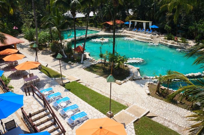 Tropical pool surrounded by lounge chairs, umbrellas, and palm trees at Hotel Chan-Kah Resort Village
