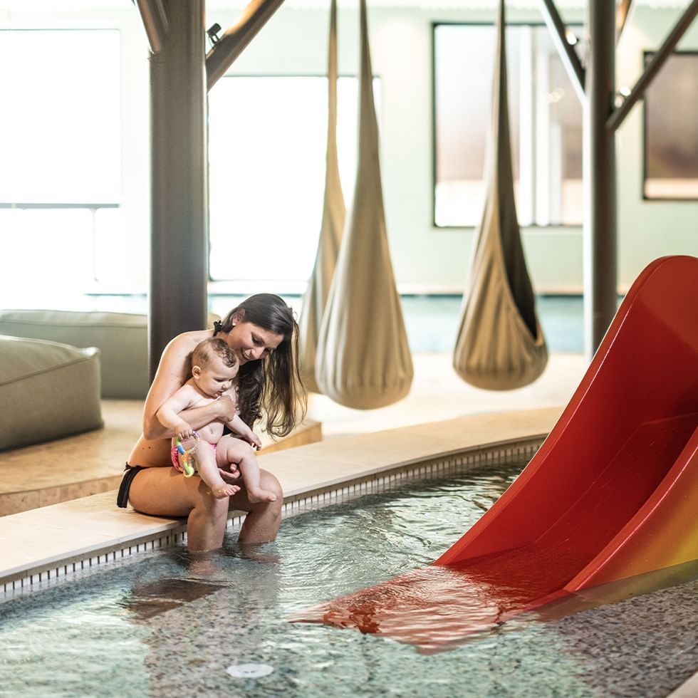 Woman holding a baby in an indoor pool with a red slide and hanging lounge chairs.