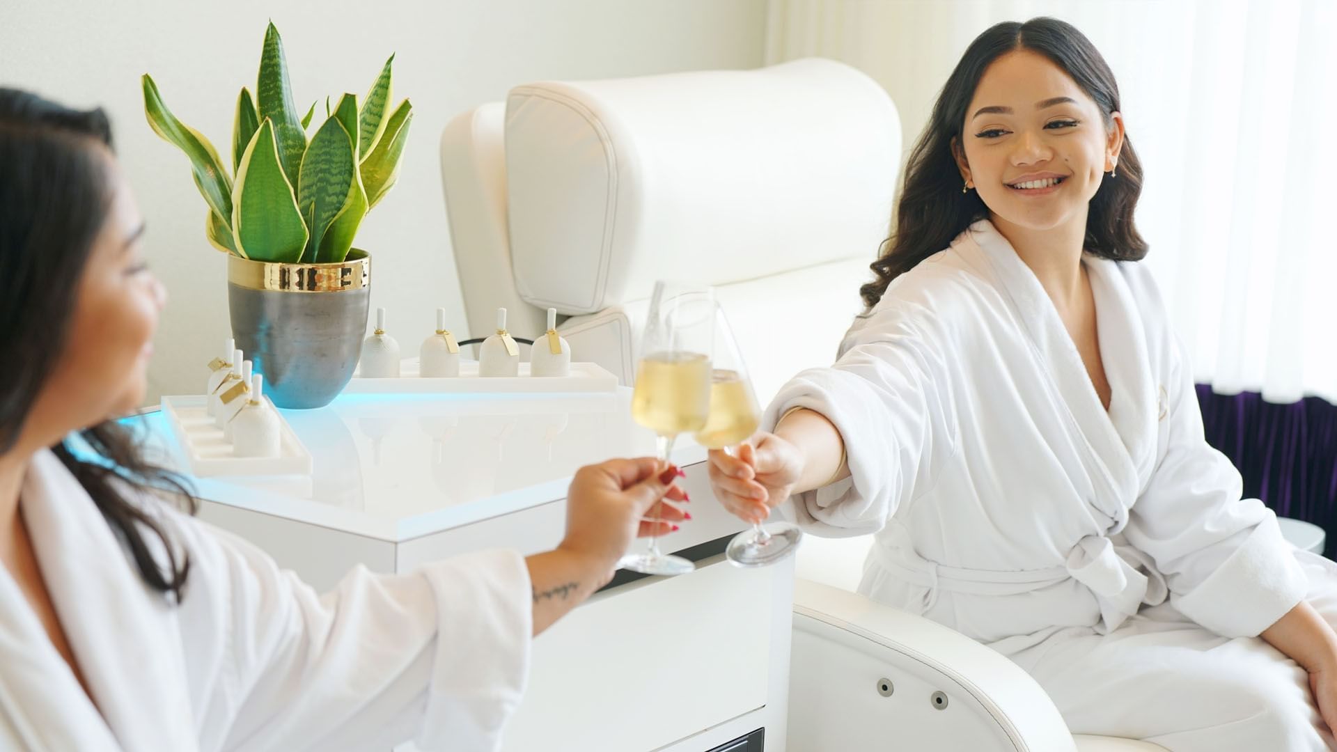 Two women in white robes clinking champagne glasses beside white treatment chairs at Guerlain Spa at Hotel X Toronto