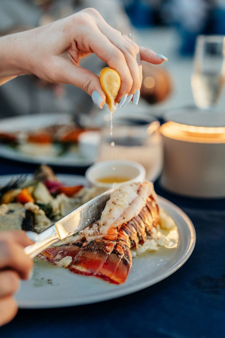 Hands holding lemon wedge over lobster on plate, squeezing juice, with knife cutting into lobster.