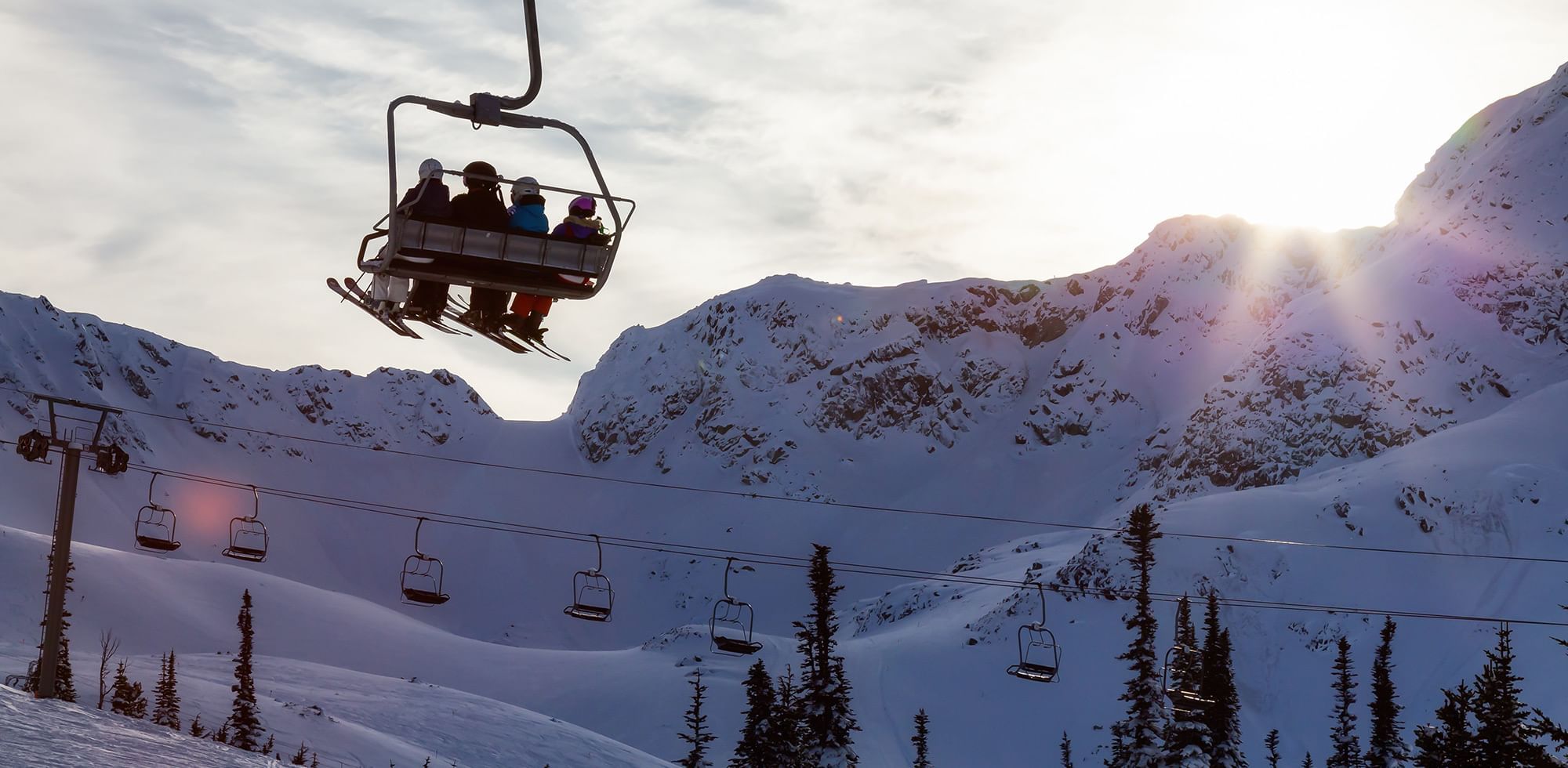 Skiers on a chairlift at sunset and surrounded by snowy mountains near Listel Whistler, a Coast Hotel