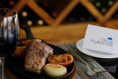 A steak with roasted onions and tomatoes on a plate next to a glass of red wine at Hotel Mykonos