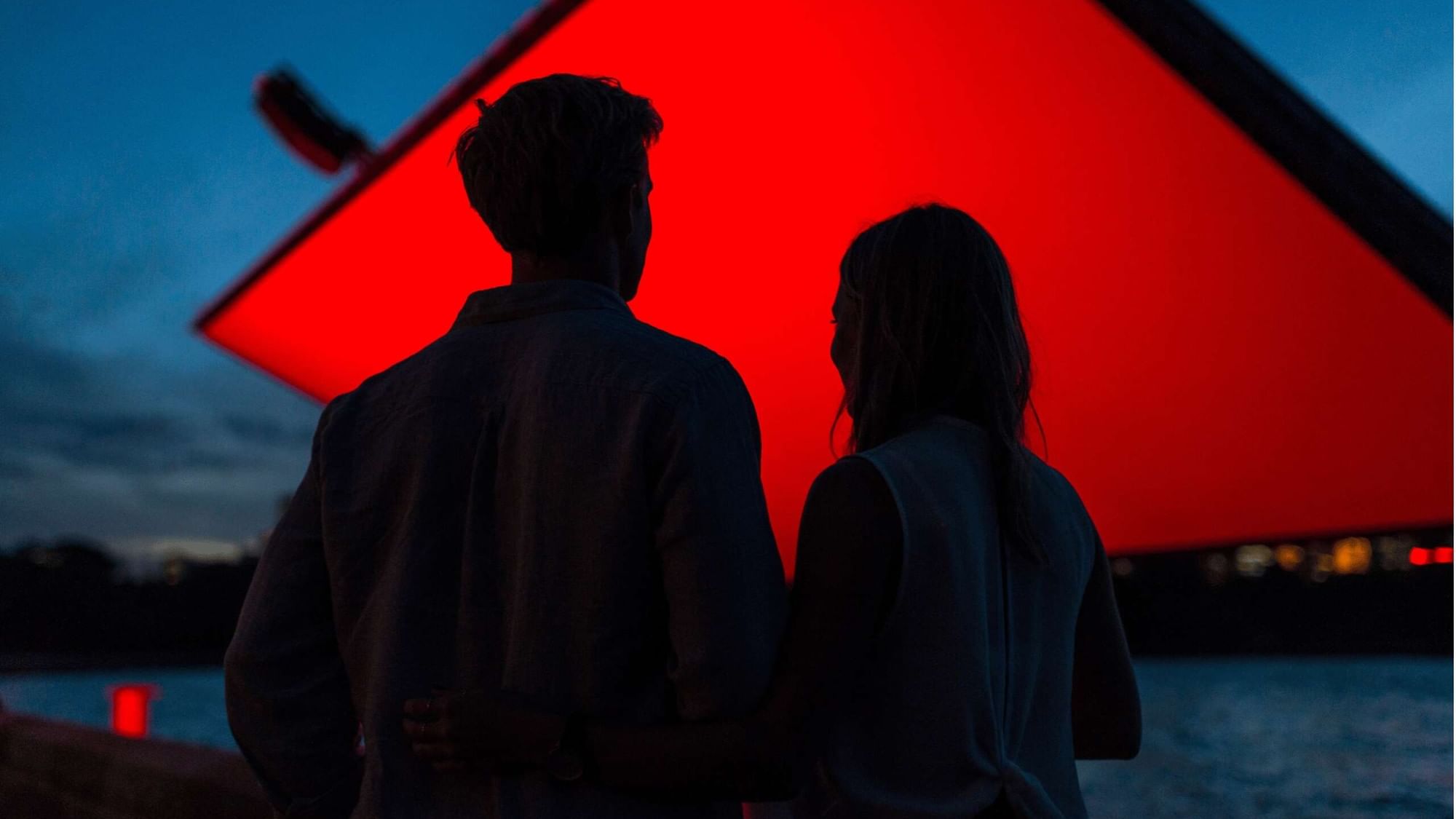 Couple watching red screen at dusk with water in background for Westpac OpenAir Cinema.