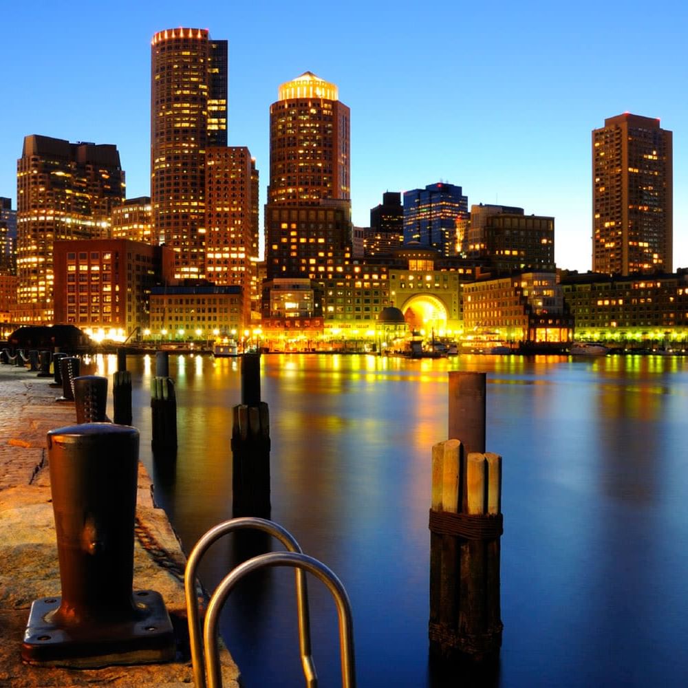 Boston skyline illuminated at dusk with harbor reflections and waterfront promenade in the foreground