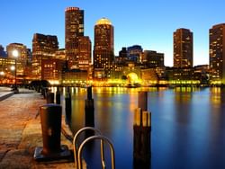 City skyline at dusk with illuminated buildings, water reflections, and a paved walkway with bollards.