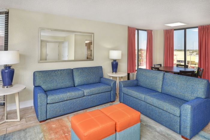 A wide view of a hotel suite living room with two blue sofas, a coffee table, and a window with a view.