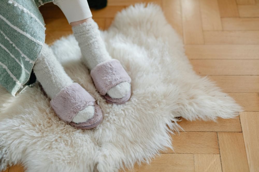 A pair of feet in fuzzy white socks and fuzzy mauve slippers on a white sheepskin rug against a chevron patterned hardwood floor.  