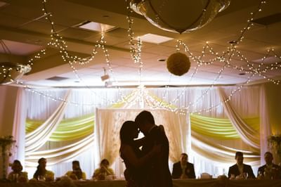 Bride and groom embracing under soft twinkling string lights, guests seated nearby at Glenmore Inn & Convention Centre