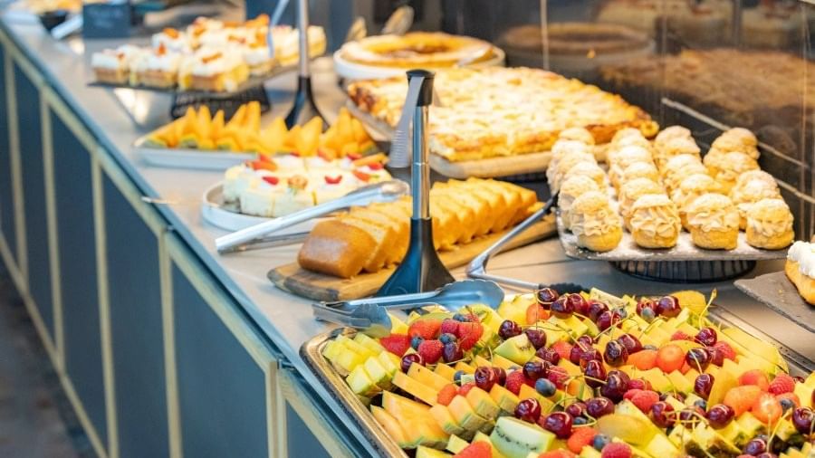 Assorted desserts and fruits on trays displayed on a buffet table with utensils and signage.