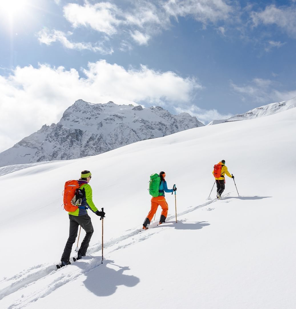Tre escursionisti con zaini colorati e bastoncini da trekking su un pendio innevato sotto un cielo parzialmente nuvoloso.