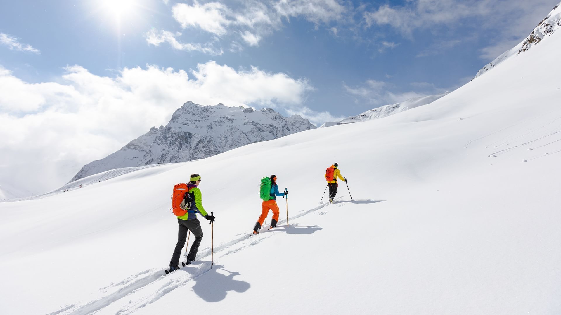 Tre escursionisti con zaini colorati e bastoncini da trekking su un pendio innevato sotto un cielo parzialmente nuvoloso.