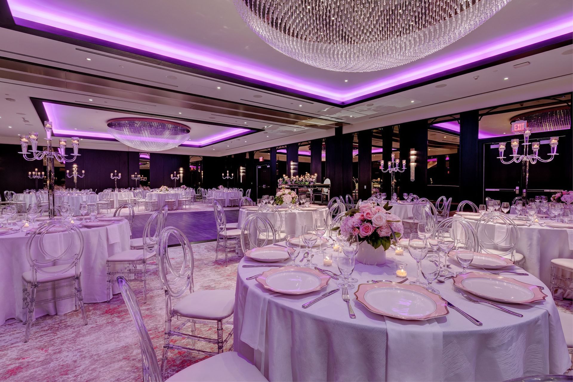 Princes Gates Ballroom set up tables with flower arrangements and clear chairs illuminated under lighting at Hotel X Toronto