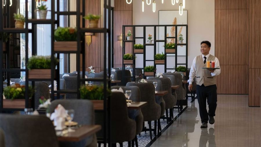 Waiter carrying colorful juices through a dining area with gray chairs and metal shelving in La Maison at Warwick Al Jubail