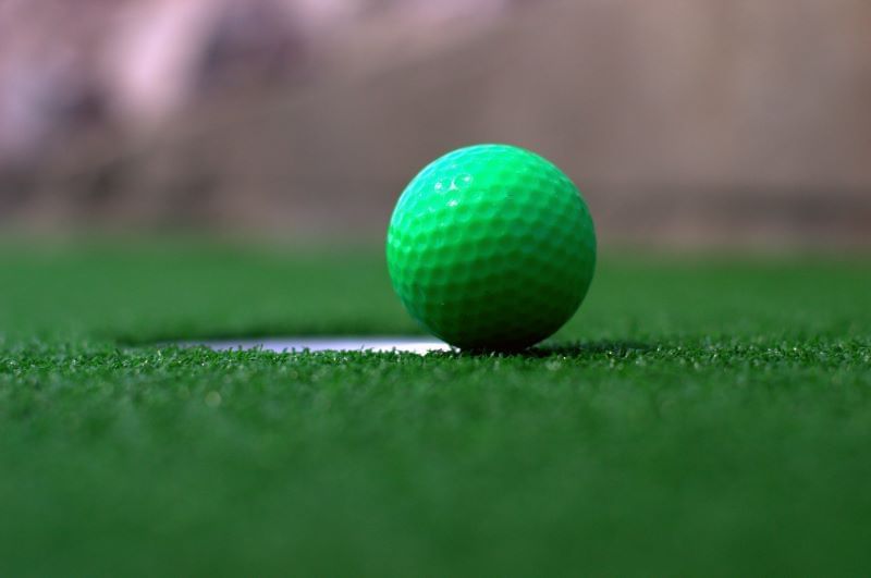 A close up image of a green golf ball on the edge of the cup (hole) on a golf course.