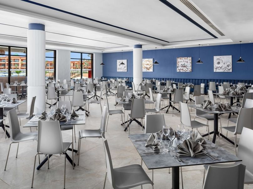 Empty restaurant with blue walls, white ceiling, gray chairs, and tables set with napkins and wine glasses.