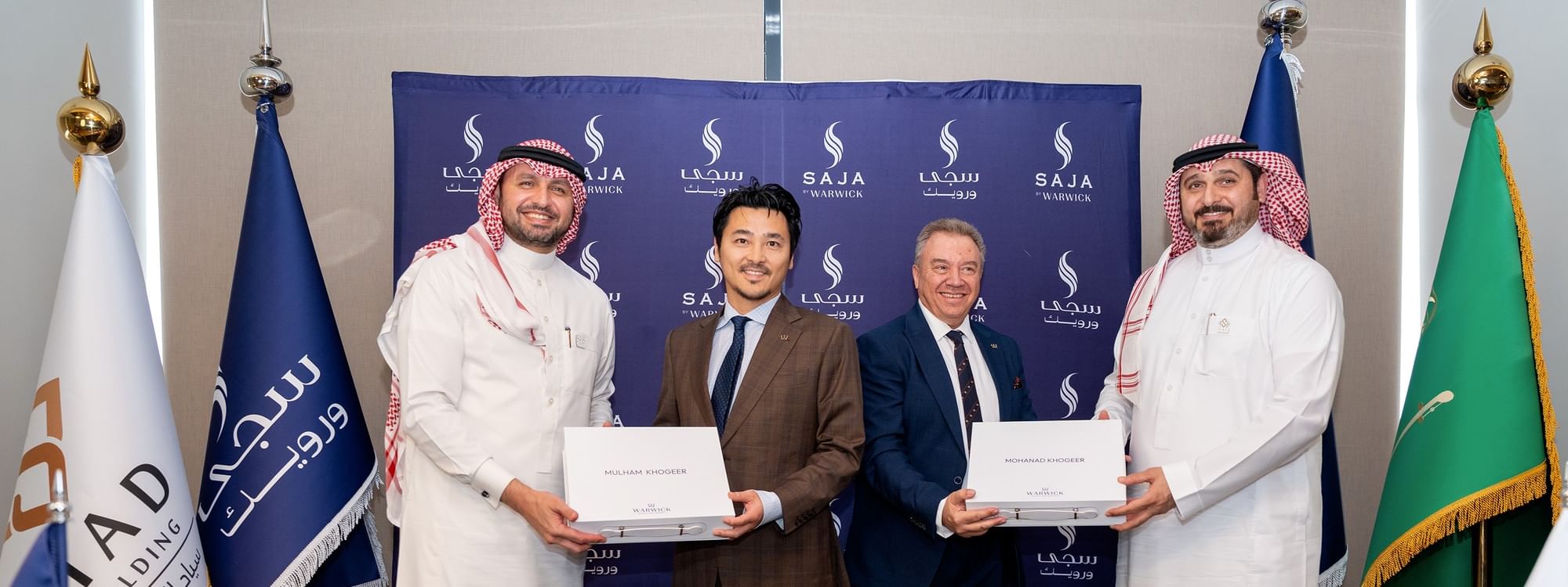 Quatre hommes en costumes devant une bannière avec des drapeaux, tenant des certificats, près de fleurs et de verres.
