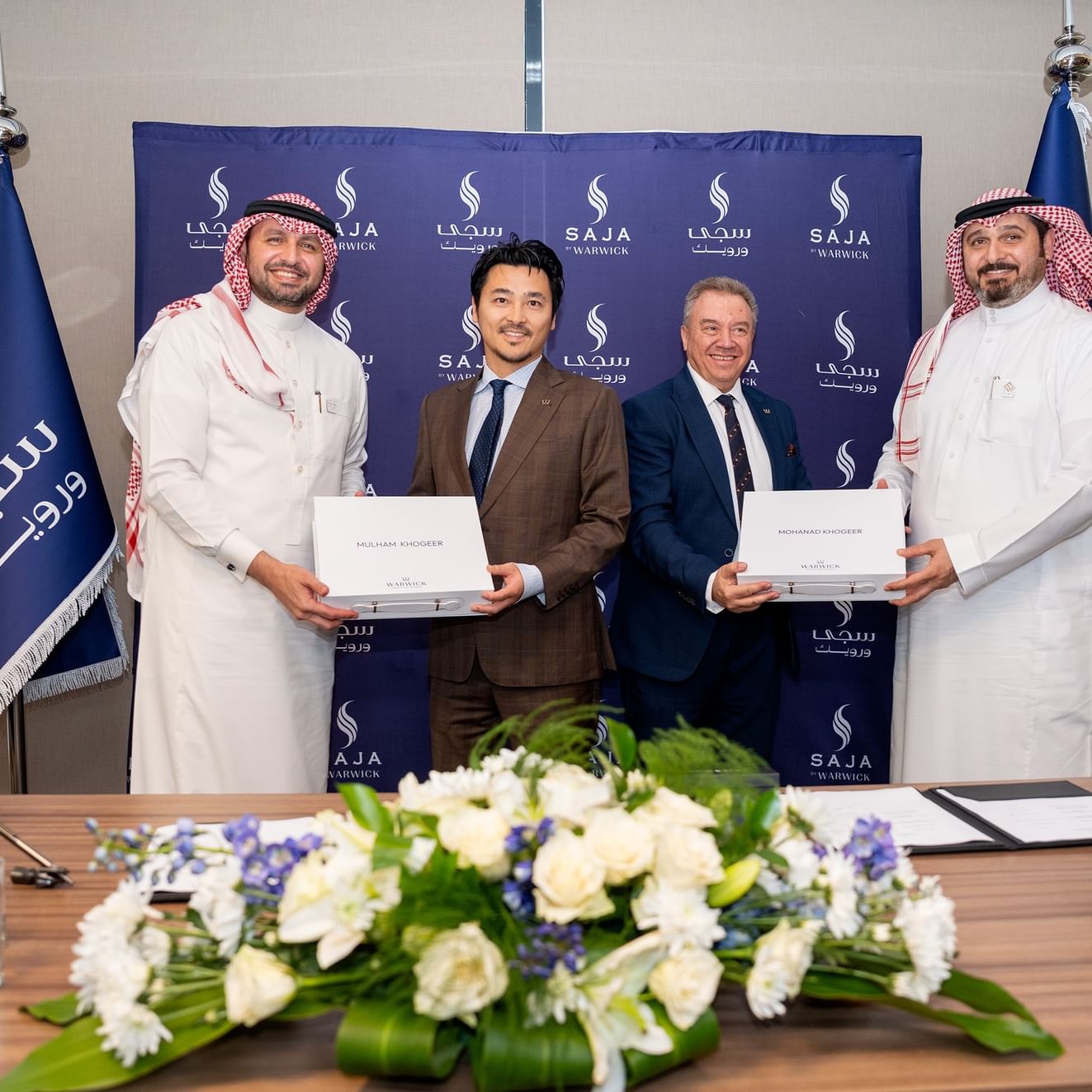 Four men in formal attire stand behind a desk with flags, holding certificates, and smiling for a photo.