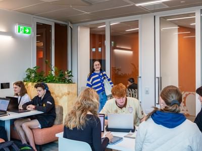 Students collaborating with laptops in a modern communal space with wooden partitions and orange accent walls at Tupuārangi.