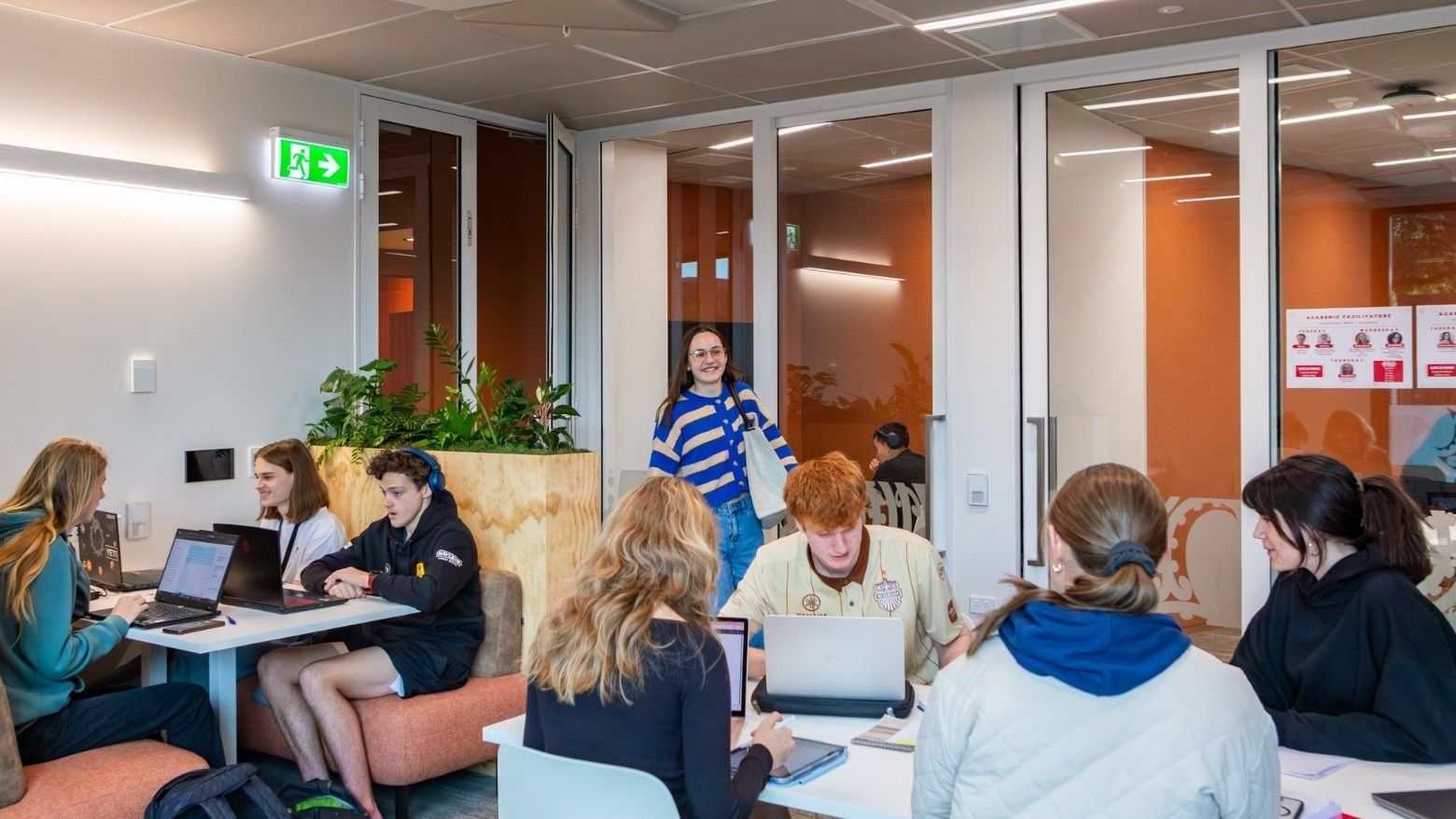 Students collaborating with laptops in a modern communal space with wooden partitions and orange accent walls at Tupuārangi.