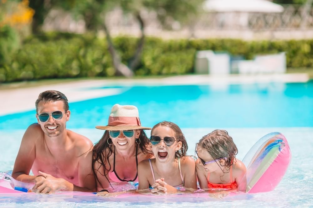 A mom, dad, and two girls, all in bathing suits and sunglasses, lean on a raft in a pool, smiling.
