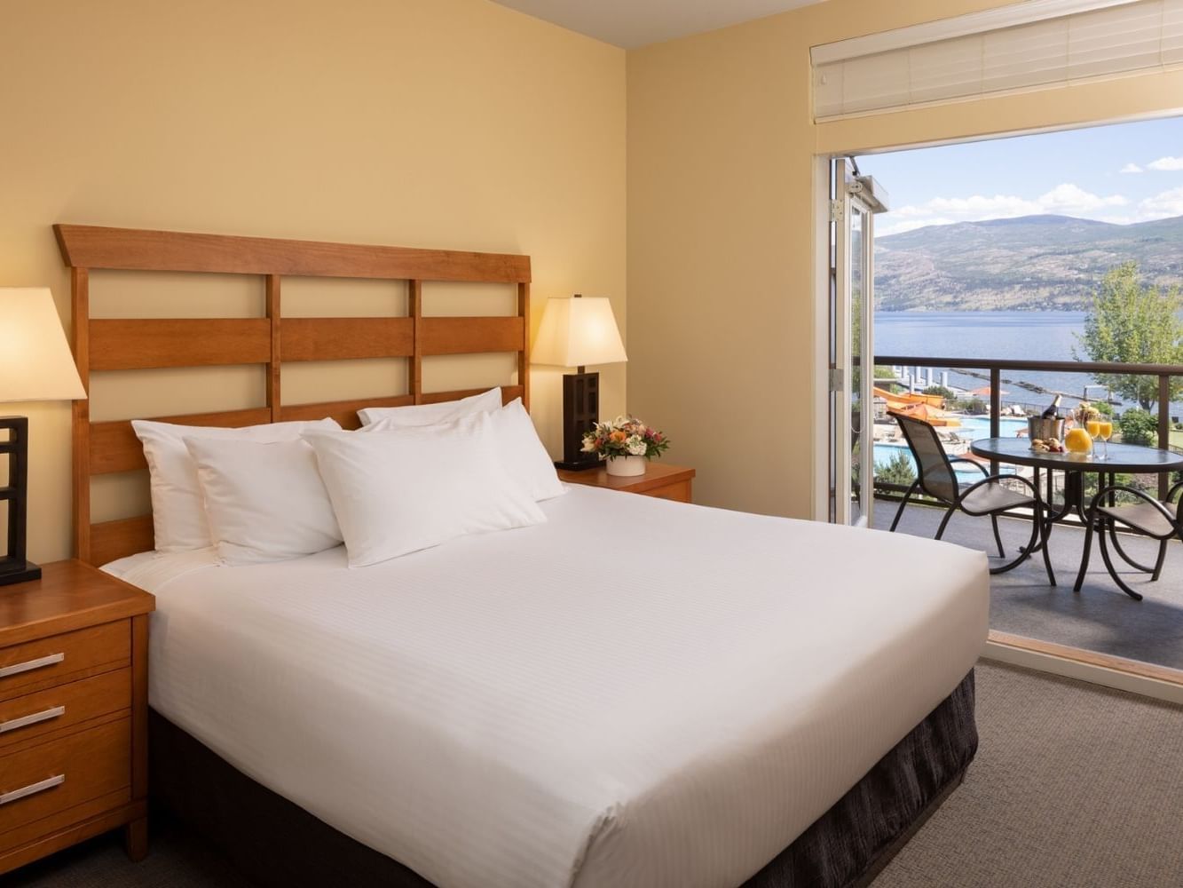 A bedroom with a bed, nightstands, lamps, and a balcony overlooking a lake and mountains.