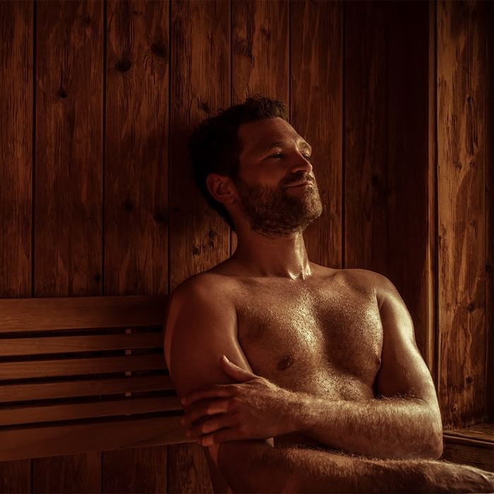 Bearded man with arms crossed, seated on a wooden bench in a wooden room, smiling.