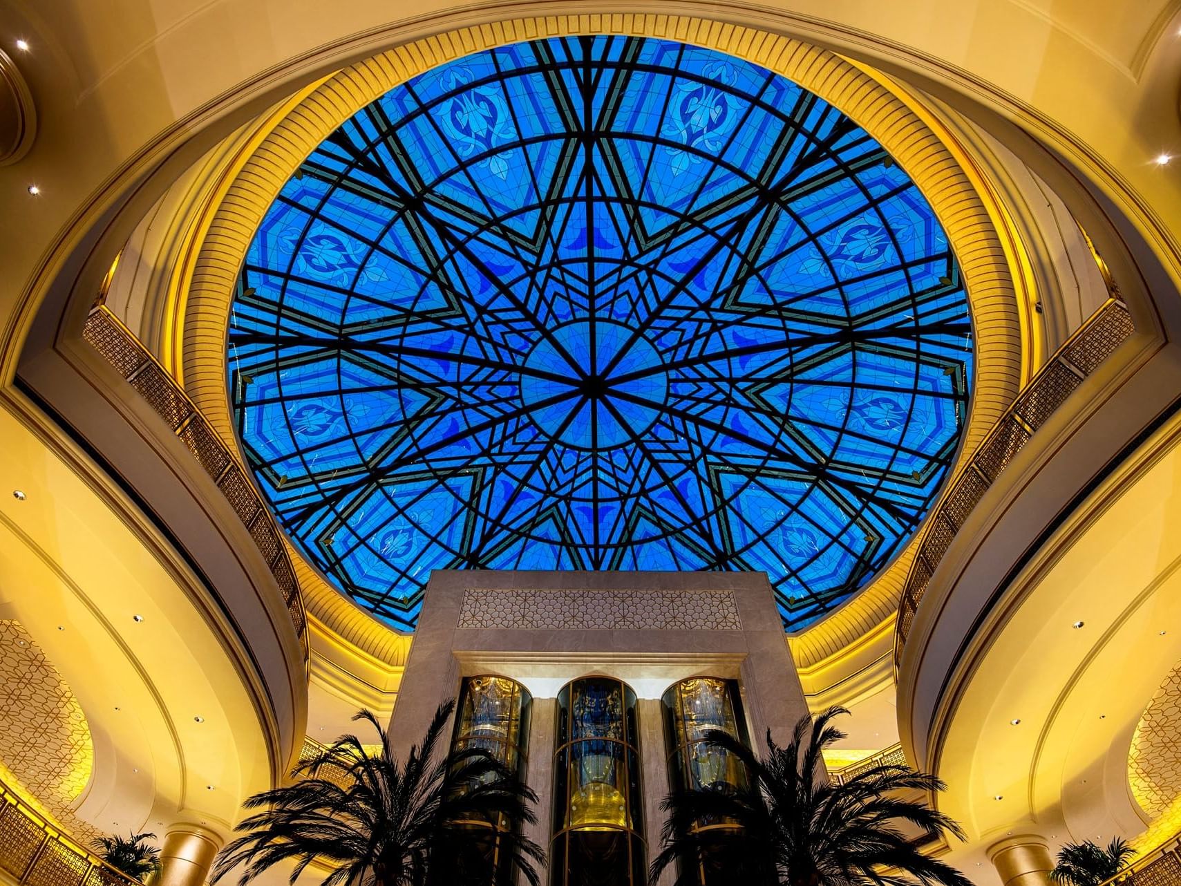 Large blue glass dome with geometric design in a luxurious interior setting with palm trees and yellow walls.