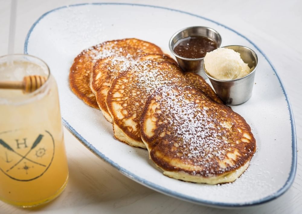 A glass with the Hampton Social logo and a honey dipper next to a plate of four golden brown pancakes on a plate with a side of butter and jam.