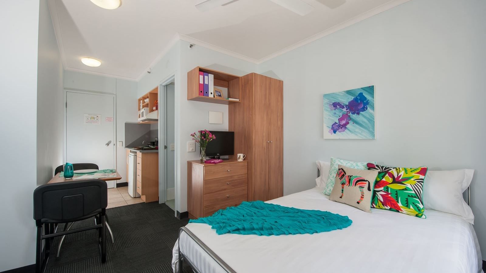 Bright studio room with white bed, colorful pillows, wooden wardrobe, kitchenette, and dining area at StudentLiving - on Margaret