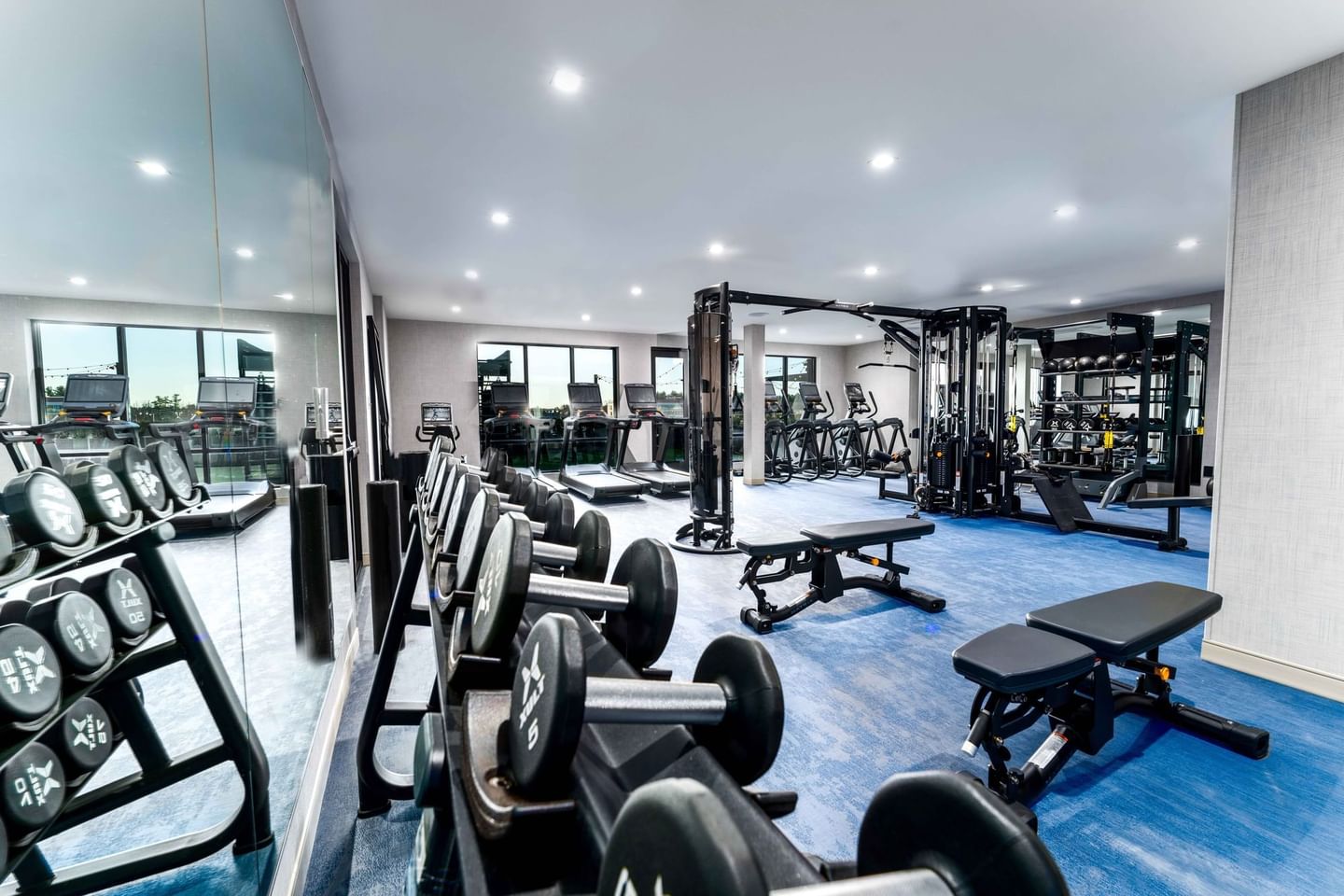 Gym with rows of dumbbells, weight machines, and cardio equipment on blue floor under a high ceiling with lights.