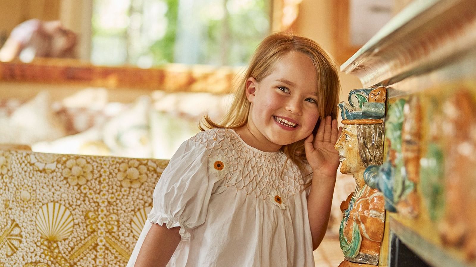 Smiling girl with a white dress standing by a colorful, tiled fireplace under natural light at the Marbella Club