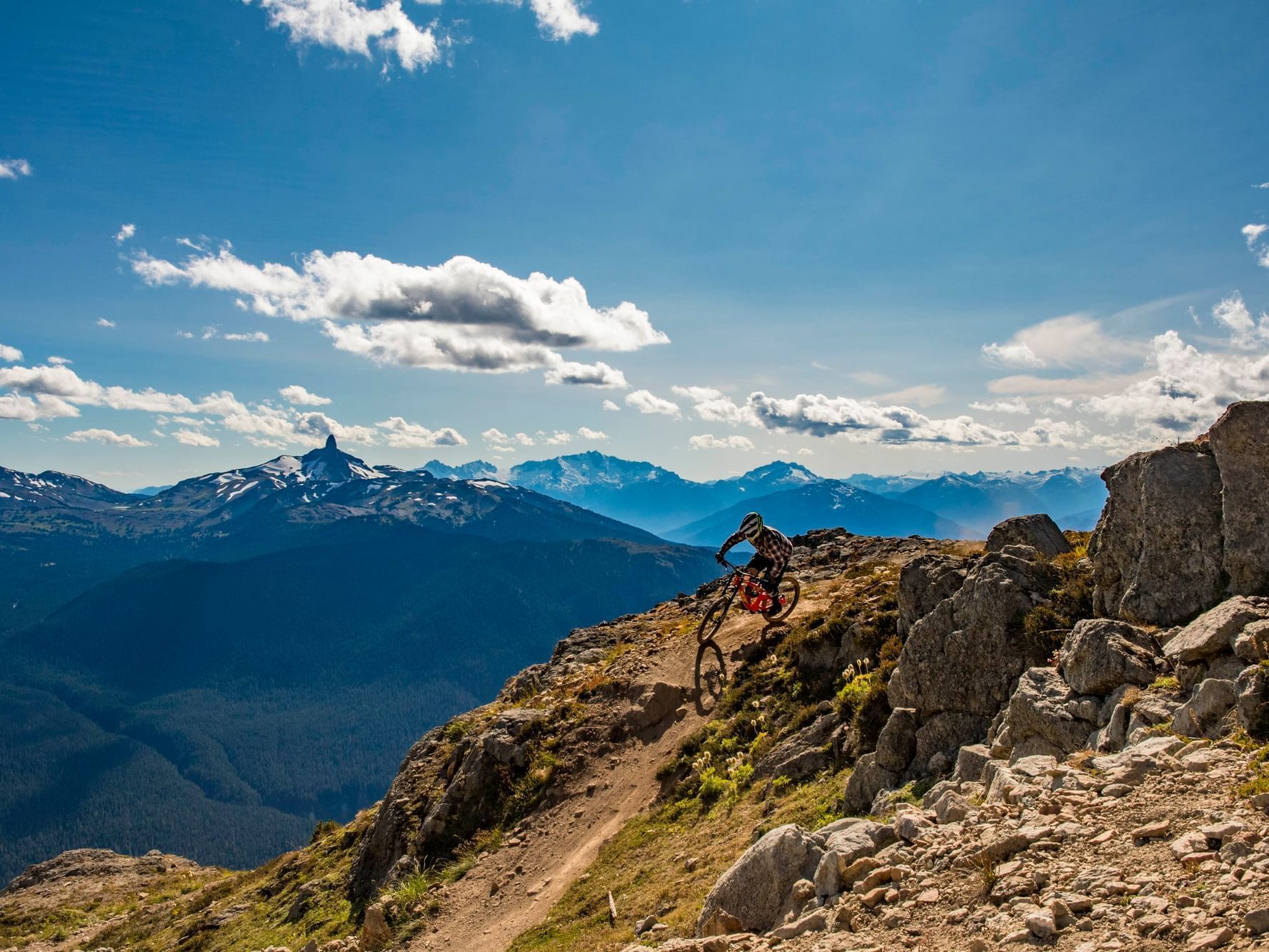 Biker descending rocky mountain trail with scenic valley and mountain range in background under a partly cloudy sky.