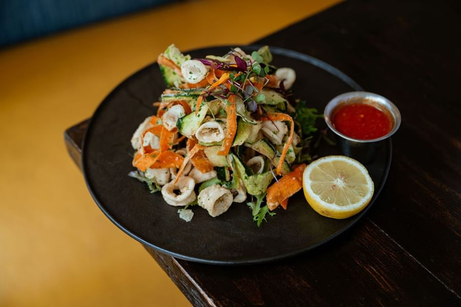 Colorful seafood salad with calamari, shredded vegetables, and lemon wedge served on a black plate with dipping sauce.