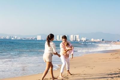 Two women walking along a sandy beach with ocean waves and coastal buildings in the background at Playa Los Arcos Puerto Vallarta