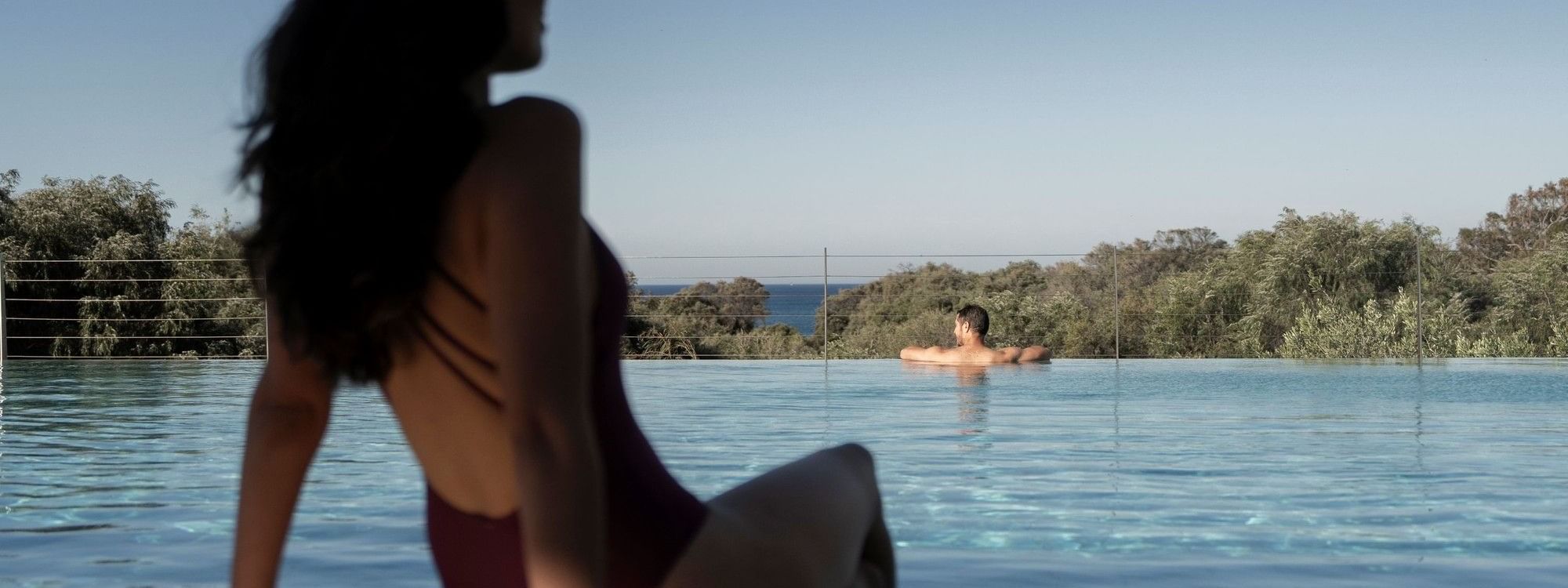 Woman in burgundy swimsuit sitting by infinity pool overlooking trees and distant ocean under clear blue sky.