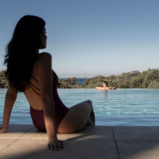 Woman in burgundy swimsuit sitting by infinity pool overlooking trees and distant ocean under clear blue sky.