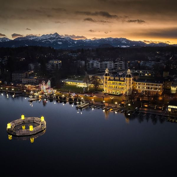 Vista aerea di una città al crepuscolo con un lago e montagne sullo sfondo.