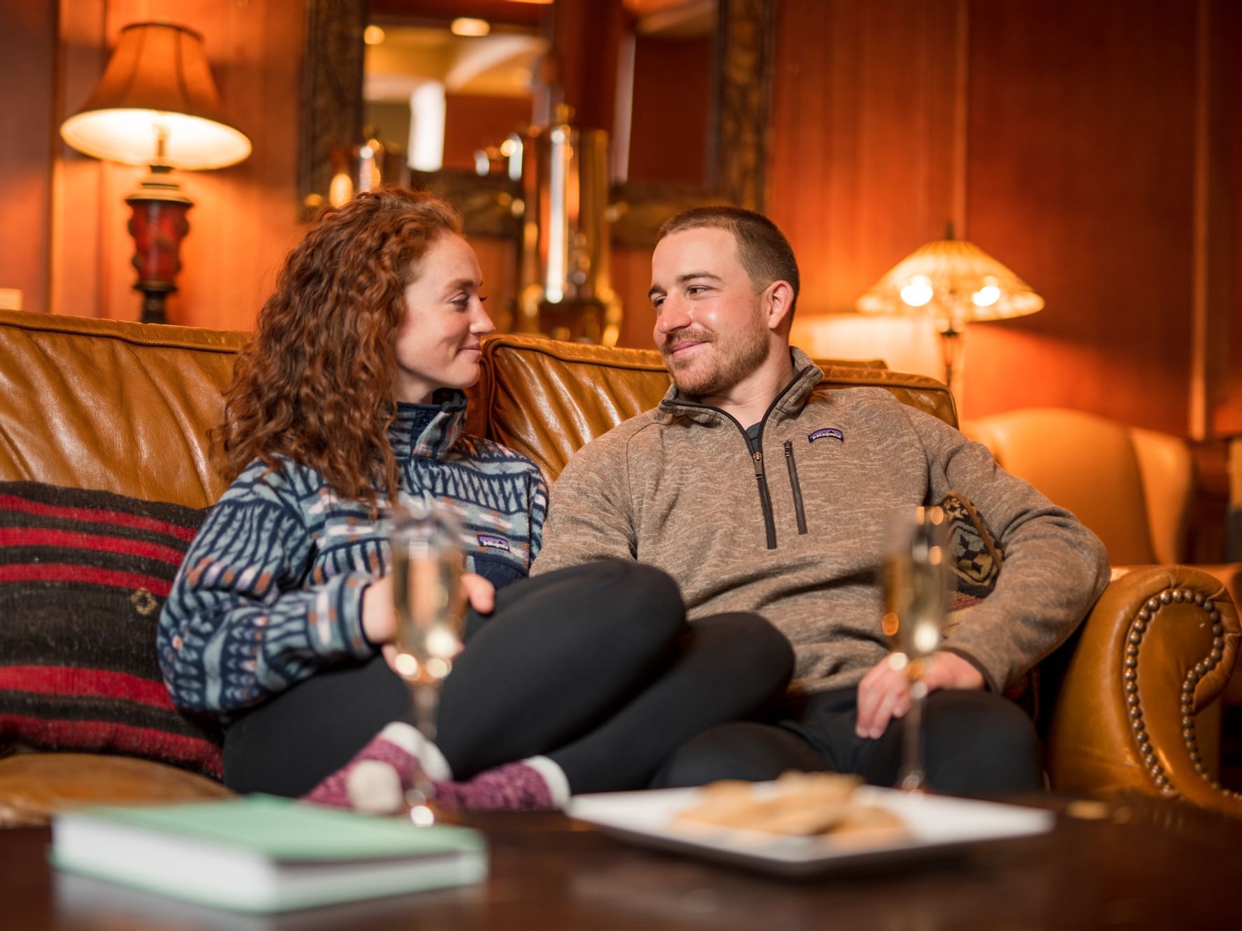 Couple sitting on a couch with champagne glasses, a plate of cookies, and a book on a coffee table.