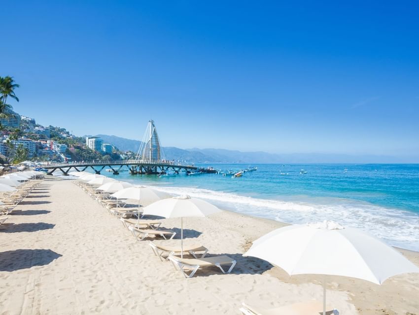 White beach umbrellas and lounge chairs on sand facing blue ocean waves and a stylish pier near Los Arcos Suites