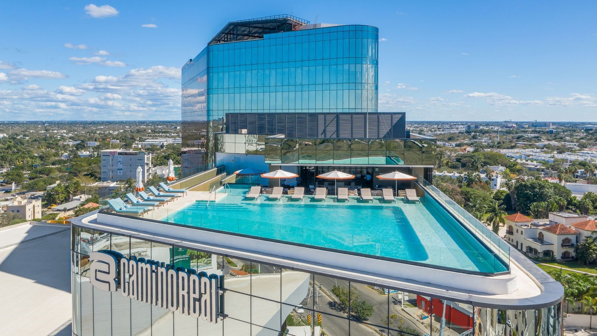 Large rooftop infinity pool featuring blue loungers and a modern glass skyscraper at Camino Real Merida