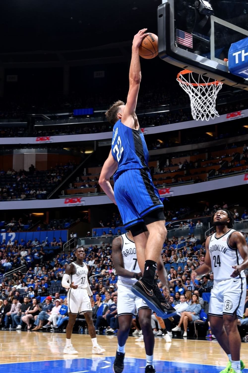 A basketball player in a blue and black uniform dunks a basketball into a net surrounded by players in white uniforms and a large arena crowd.