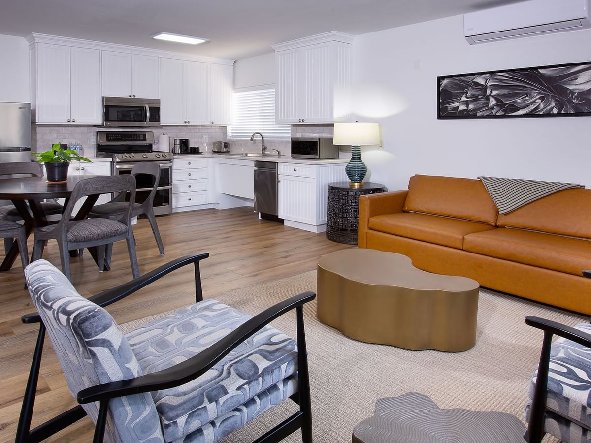 A modern hotel living room with an orange couch, coffee table, dining set, and kitchen area.