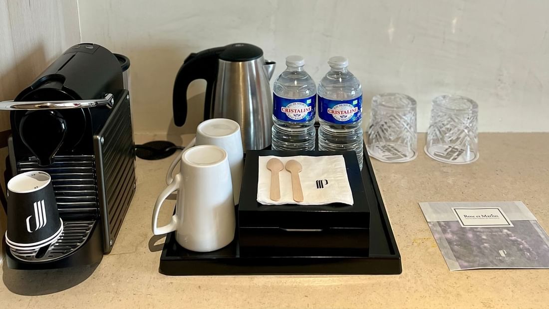 Coffee machine, kettle, water bottles, cups, spoons, and a paper on a countertop.