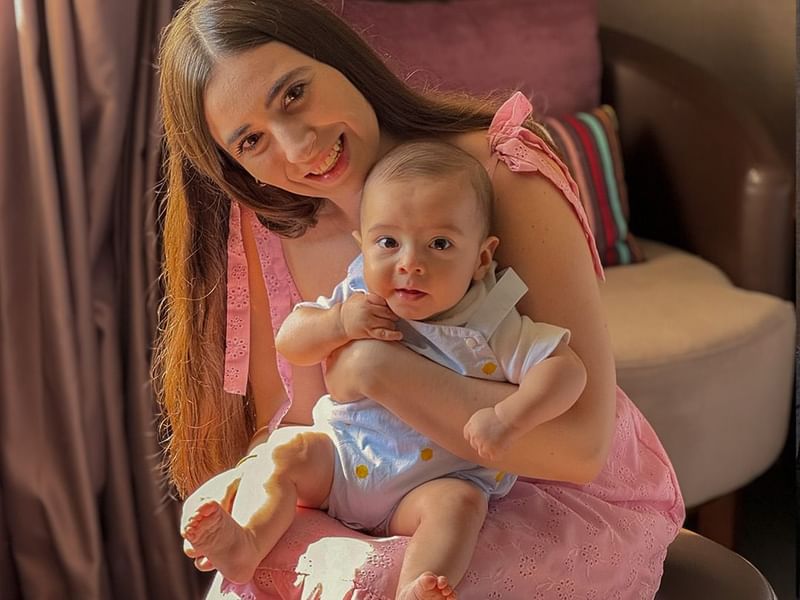Woman in pink dress smiles holding a baby on her lap in a cozy, softly lit room at Fiesta Americana Travelty