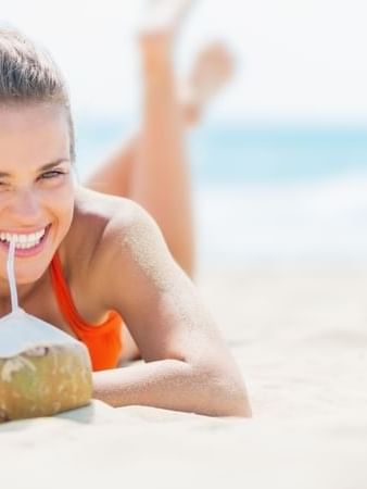 Smiling guest in an orange swimsuit enjoying a fresh coconut drink on the white sand at TokaToka Resort Nadi Fiji
