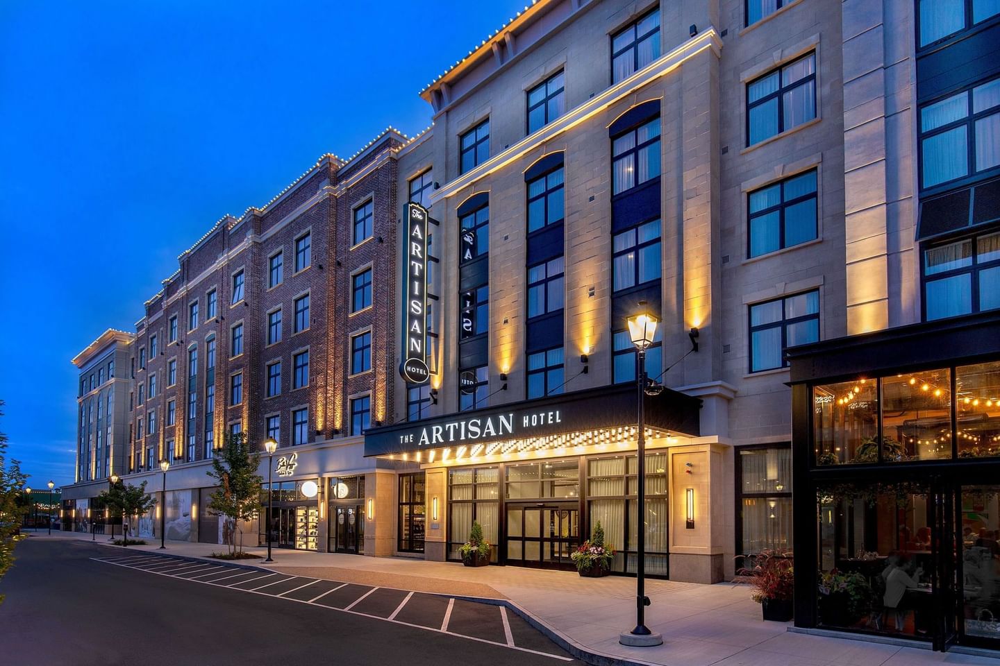The Artisan Hotel with illuminated signage and large windows at dusk, next to a restaurant with lit interior.