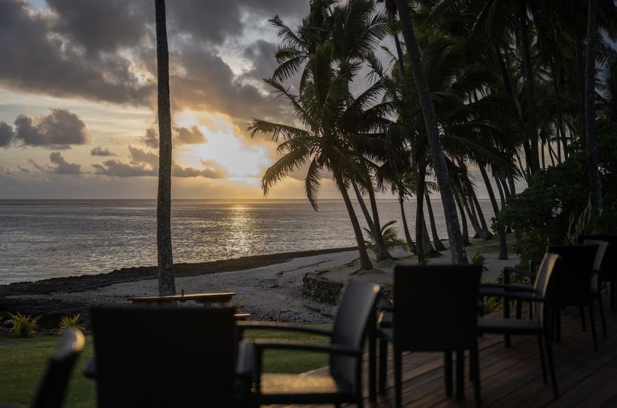 Wicker chairs by a wooden deck under tall palm trees during a golden sunset near the ocean at Tambua Sands Beach Resort