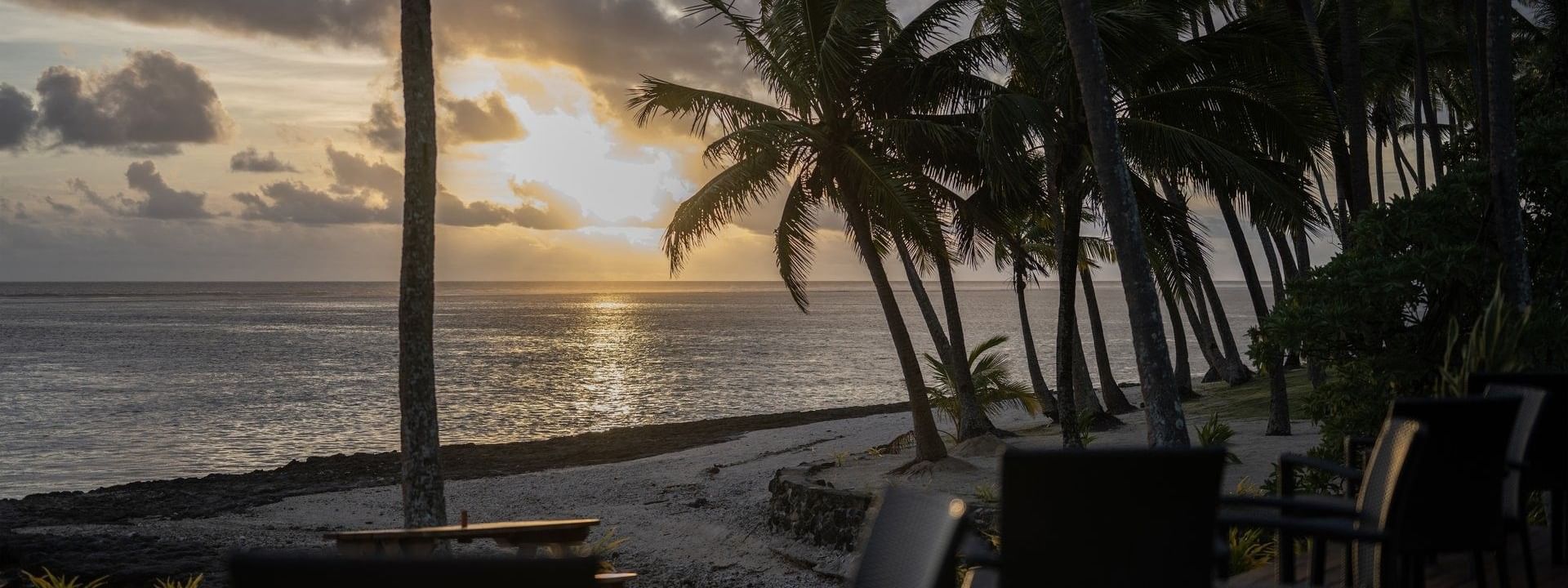 Dining chairs on a wooden deck by palm trees under a golden sunset near the beach at Warwick Hotels & Resorts