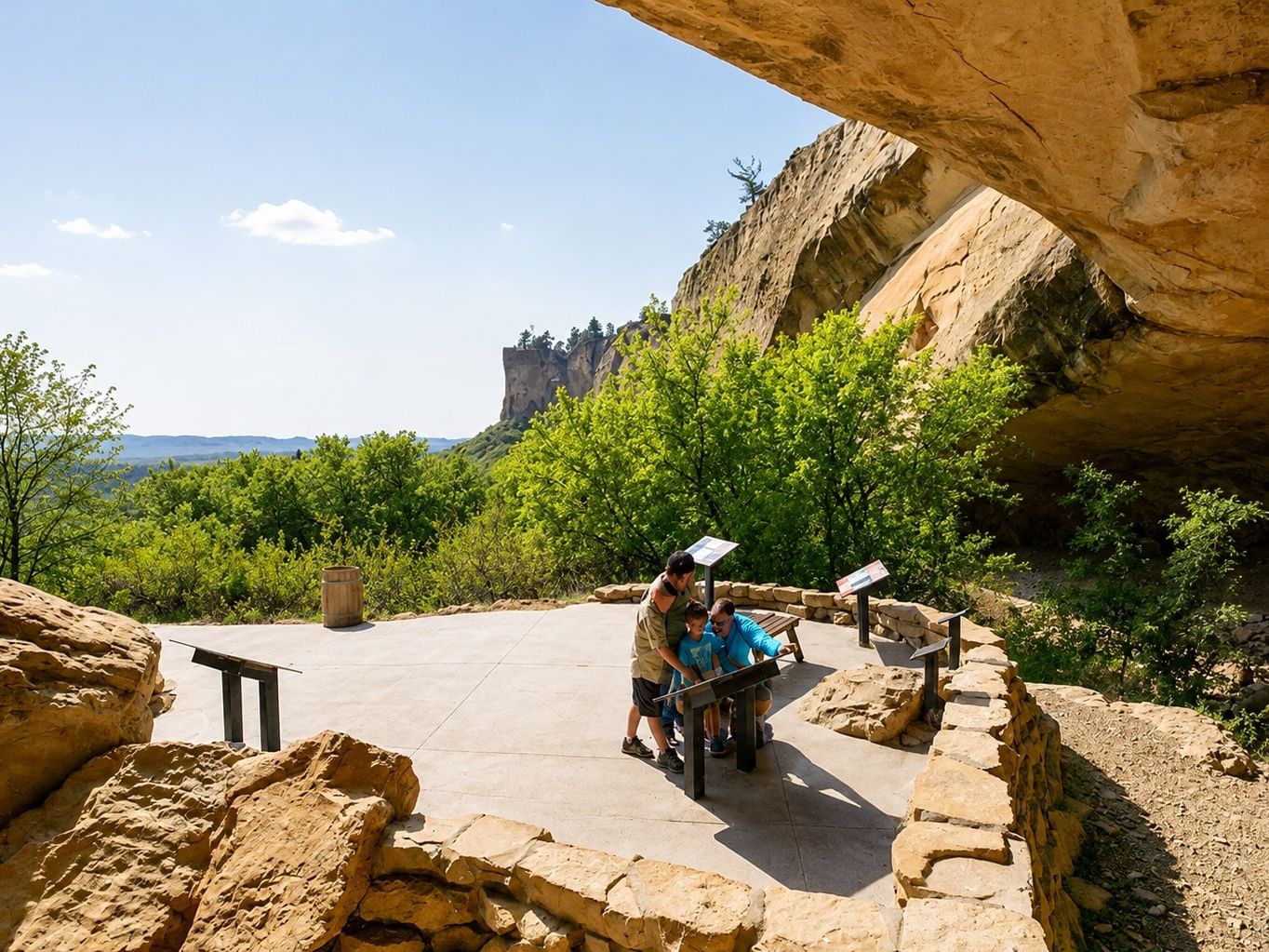 Visitors reading interpretive signs beneath a sandstone overhang with distant rock formations at Pictograph Cave State Park.